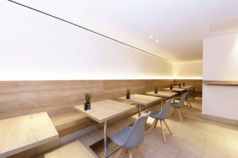 Een modern café met houten tafels en grijze stoelen, langs een met hout betimmerde muur met kleine planten op elke tafel. Rustige sfeer.