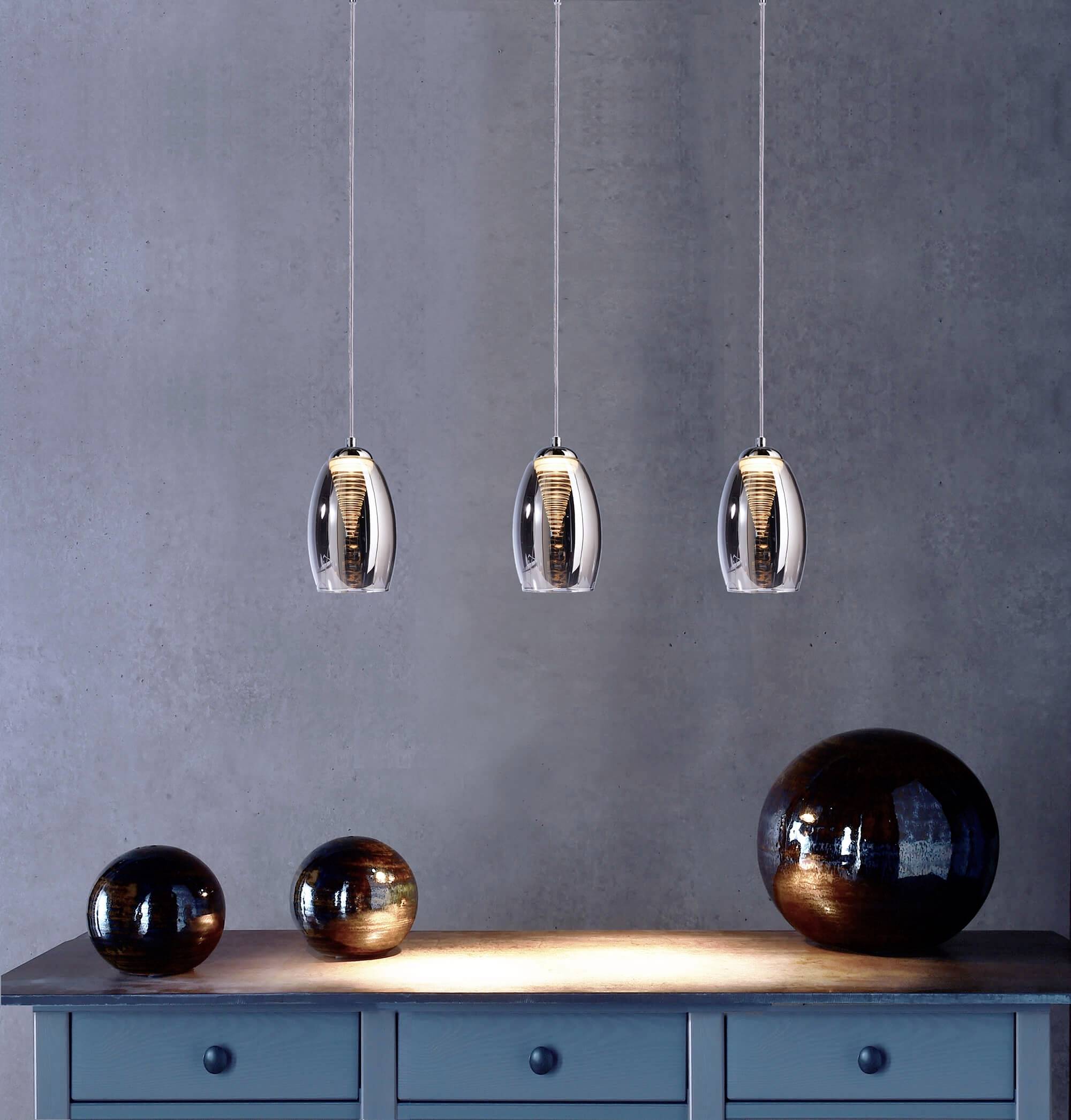Drie hangende designlampen boven een blauwe tafel met drie houten decoratieve bollen in verschillende formaten op een grijze achtergrond.