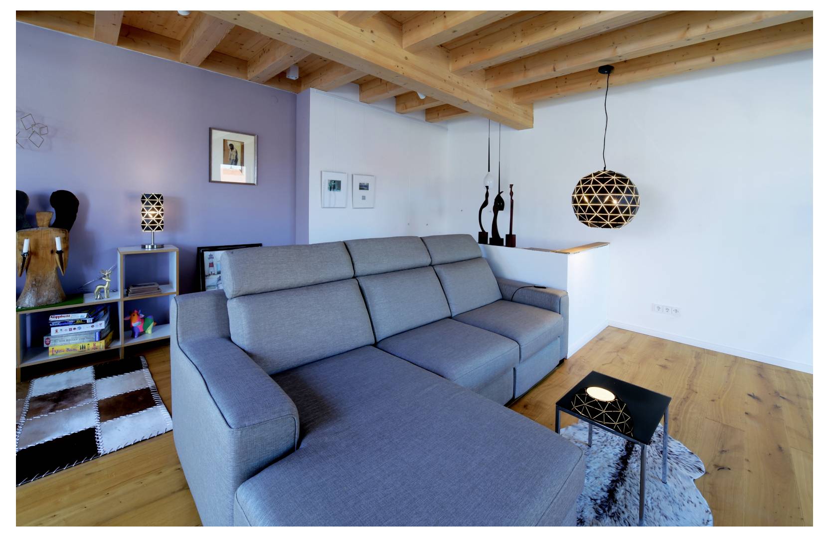 Moderne woonkamer met een grijze hoekbank, houten plafondbalken, zwarte hanglampen, een zijplank en een klein salontafeltje op een vloerkleed.