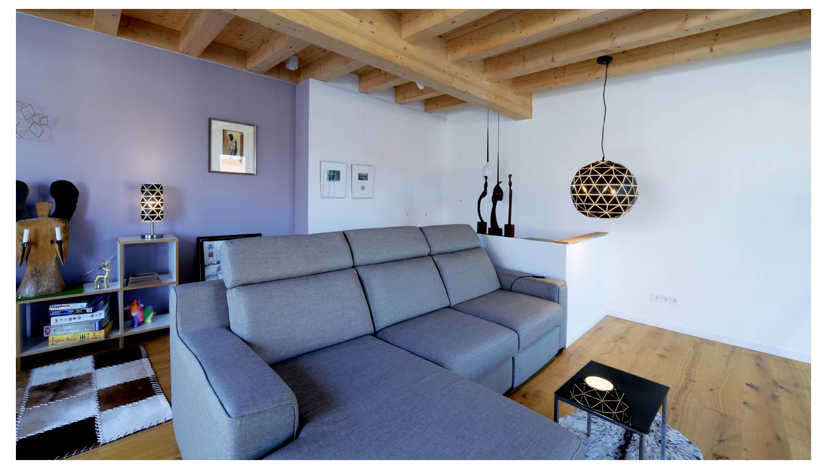 Moderne woonkamer met een grijze hoekbank, houten plafondbalken, een kleine zwarte tafel, open kasten en een hanglamp in een helder, minimalistisch interieur.
