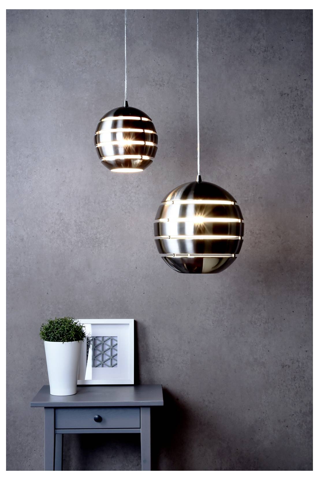 Twee moderne hanglampen hangen boven een klein bijzettafeltje met een potplant en ingelijste kunst tegen een grijze muur.
