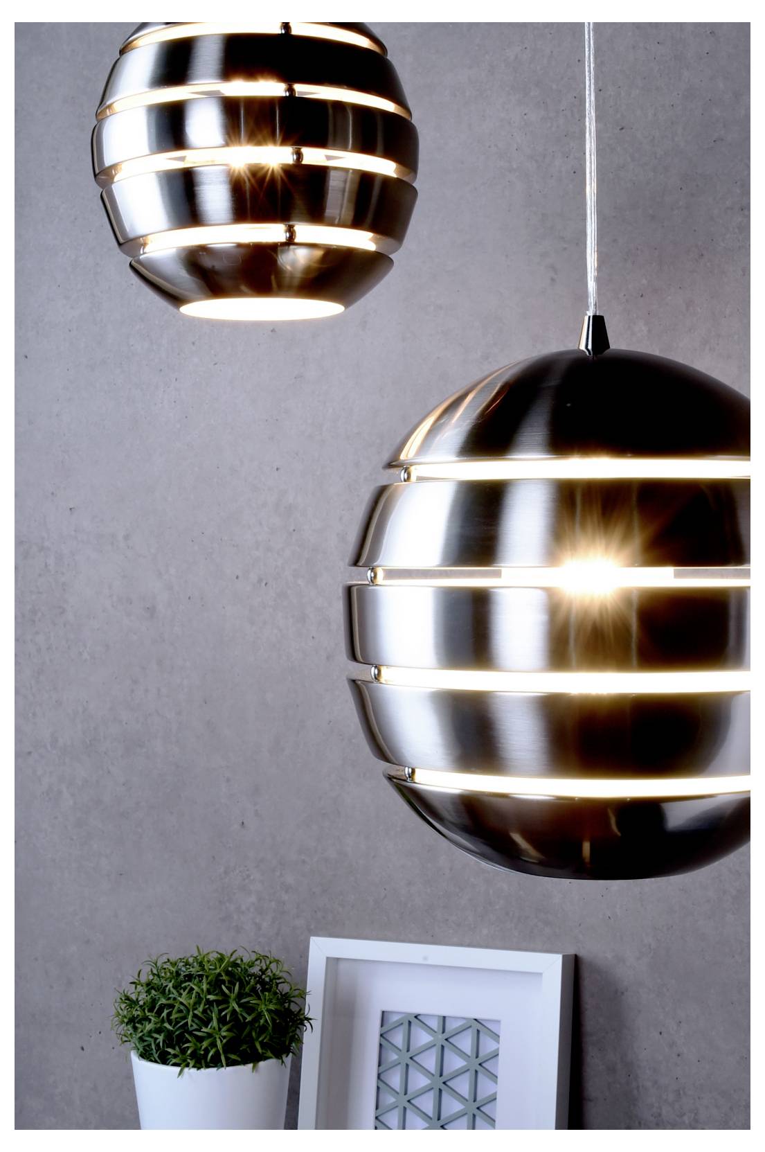 Twee moderne metalen hanglampen hangen tegen een grijze muur boven een kleine potplant en ingelijst kunstwerk.