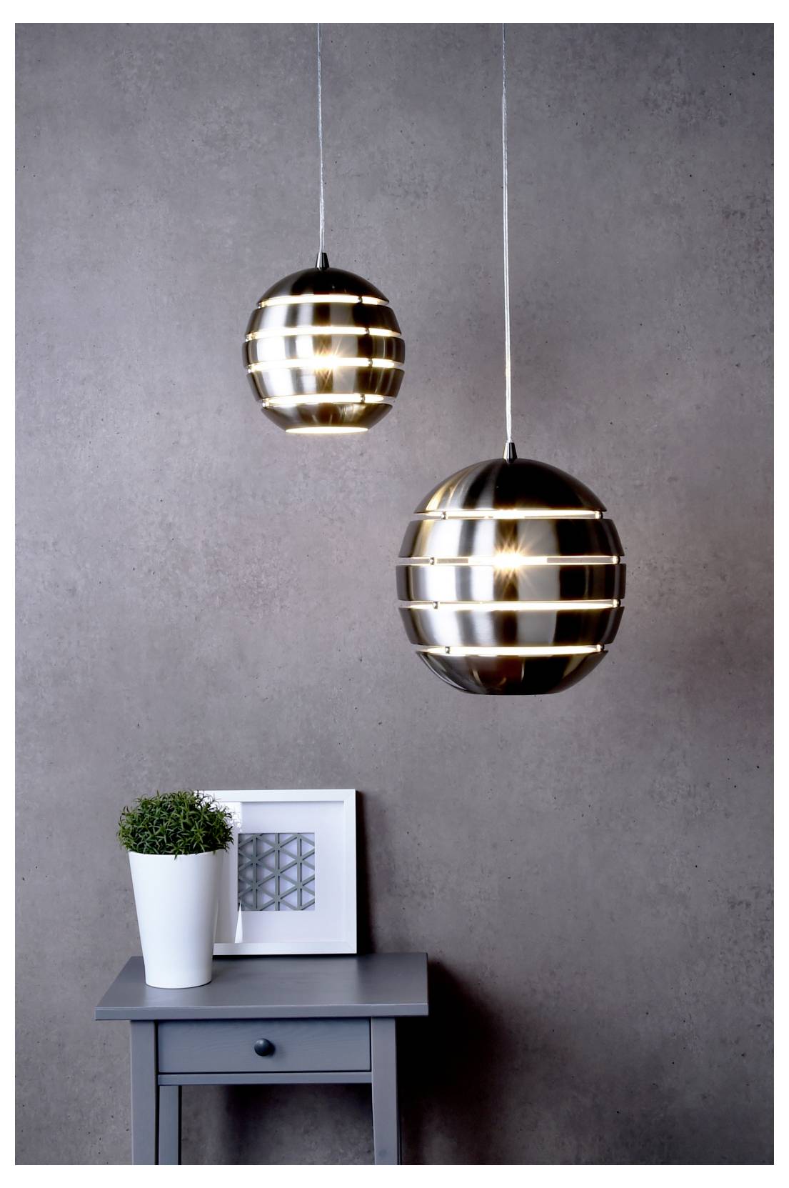 Twee hanglampen boven een klein grijs tafeltje met een geplante pot en ingelijste kunst tegen een effen grijze muur.