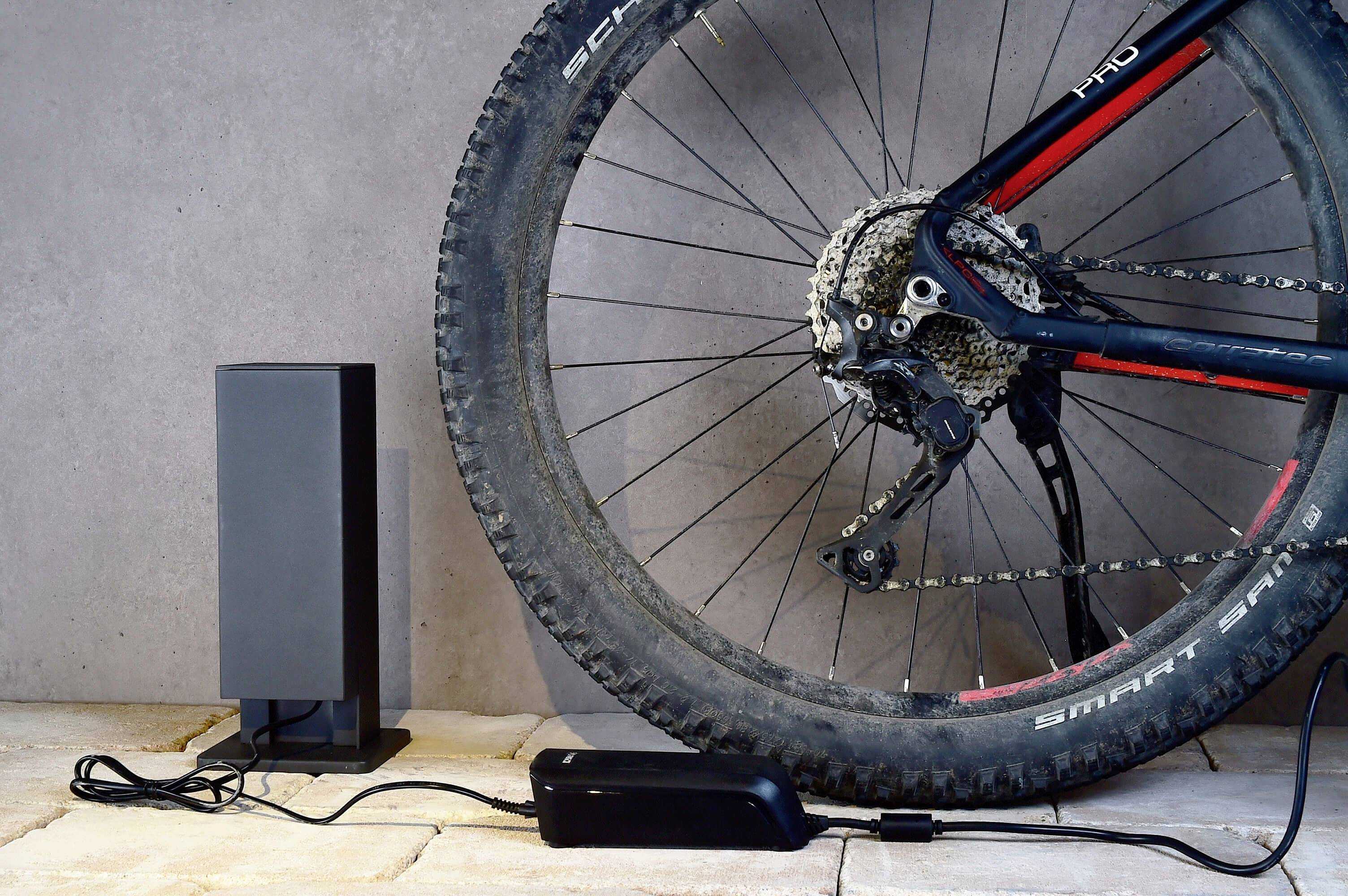 Een e-bike met aangesloten oplader staat tegen een grijze muur. De oplader is aangesloten op een stopcontact.