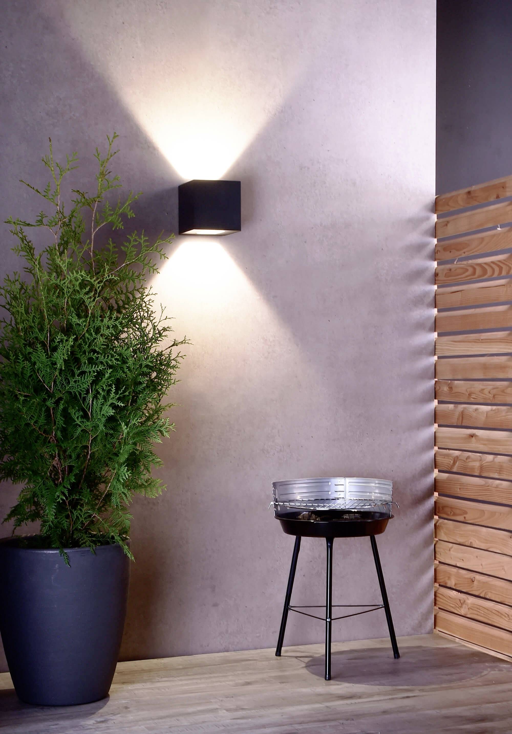 Een moderne ruimte met een verlichte wandlamp, een hoge, groene kamerplant links en een kleine, ronde barbecue op een standaard rechts.