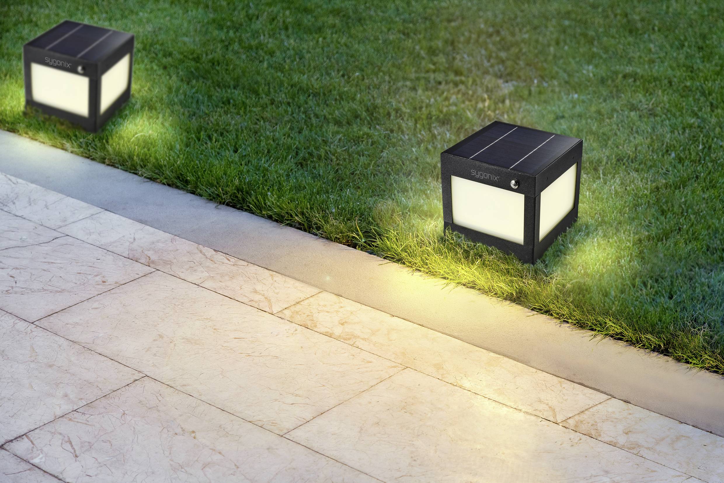 Twee op zonne-energie werkende lampen verlichten een tuinpad. De lampen hebben een rechthoekige vorm en staan op een grasveld naast het pad.
