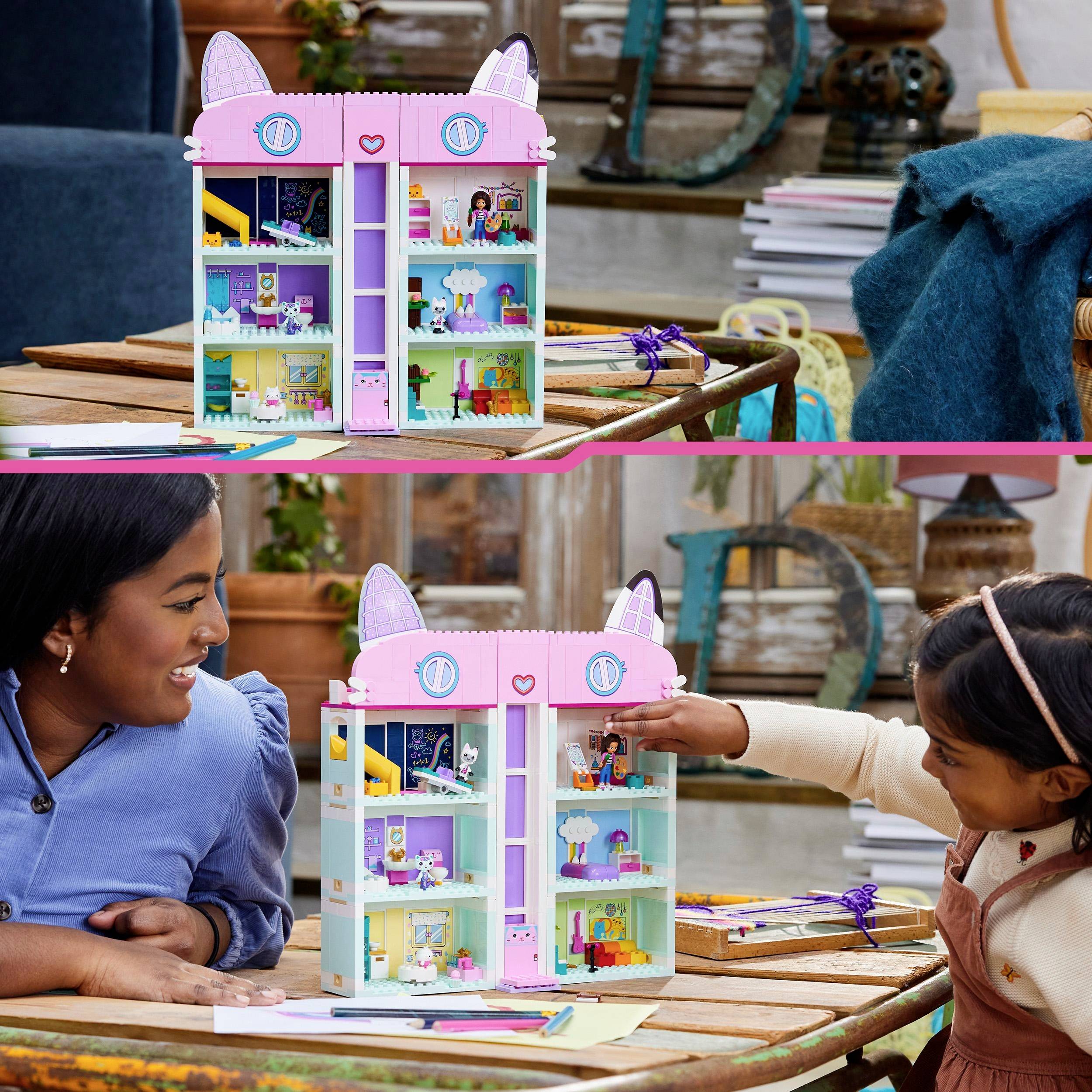 Een kind en een volwassene zitten aan een tafel en spelen met een kleurrijk, opvouwbaar poppenhuis. Het kind wijst naar figuren.