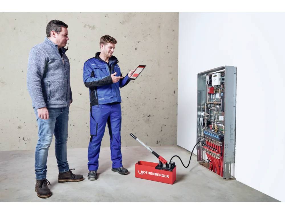 Een werknemer in blauwe overall bedient een Rothenberger pijptestapparaat, terwijl een andere persoon toekijkt. Ze inspecteren een mechanisch paneel.