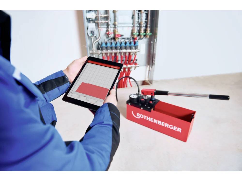 Een persoon gebruikt een tablet om drukgegevens te controleren naast een rode Rothenberger druktest-apparaat bij een paneel met meerdere kleppen.