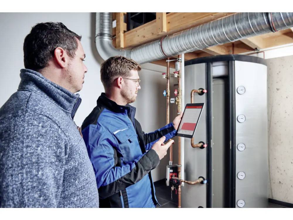 Twee technici inspecteren industriële apparatuur. Een van hen houdt een tablet vast waarop gegevens worden weergegeven, terwijl de ander toekijkt. Buizen en meters zijn zichtbaar.