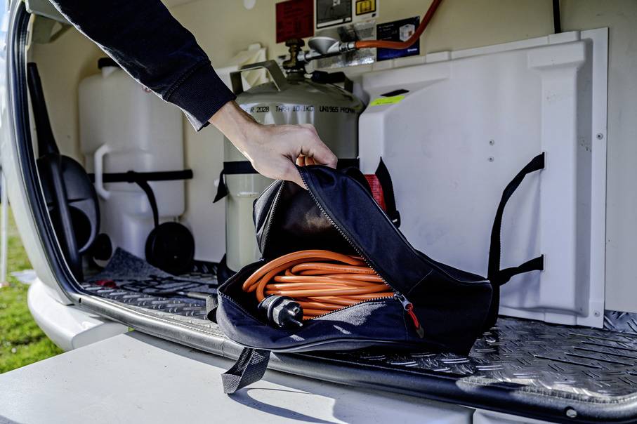 Een persoon bergt een oranje kabel op in een tas in de laadruimte van een camper. Op de achtergrond zijn watertanks zichtbaar.