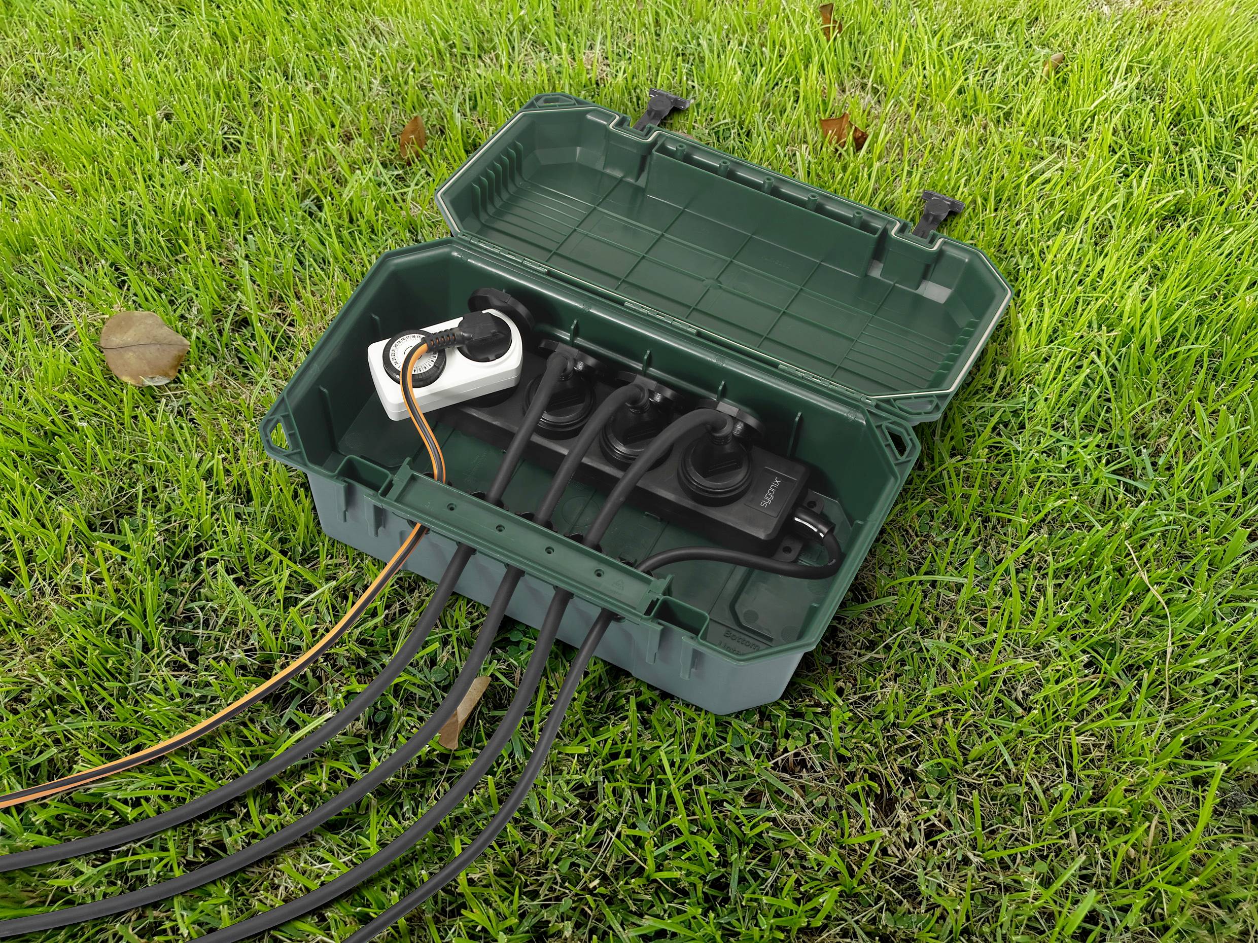 Een groene, geopende kabelbox staat op gras en bevat een stekkerdoos met meerdere aangesloten kabels.