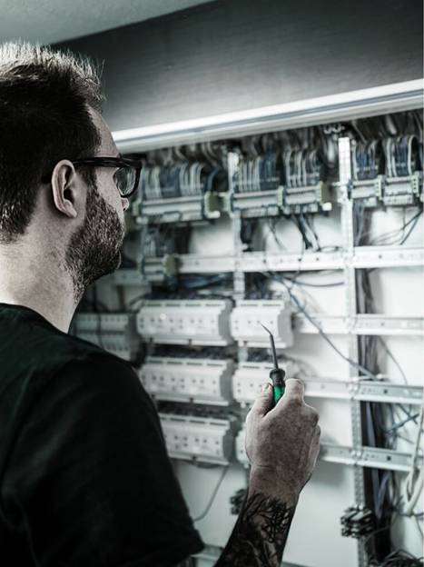 Een persoon die een schroevendraaier vasthoudt, inspecteert een groot elektrisch paneel met talrijke draden en schakelaars, wat wijst op een onderhoudstaak.