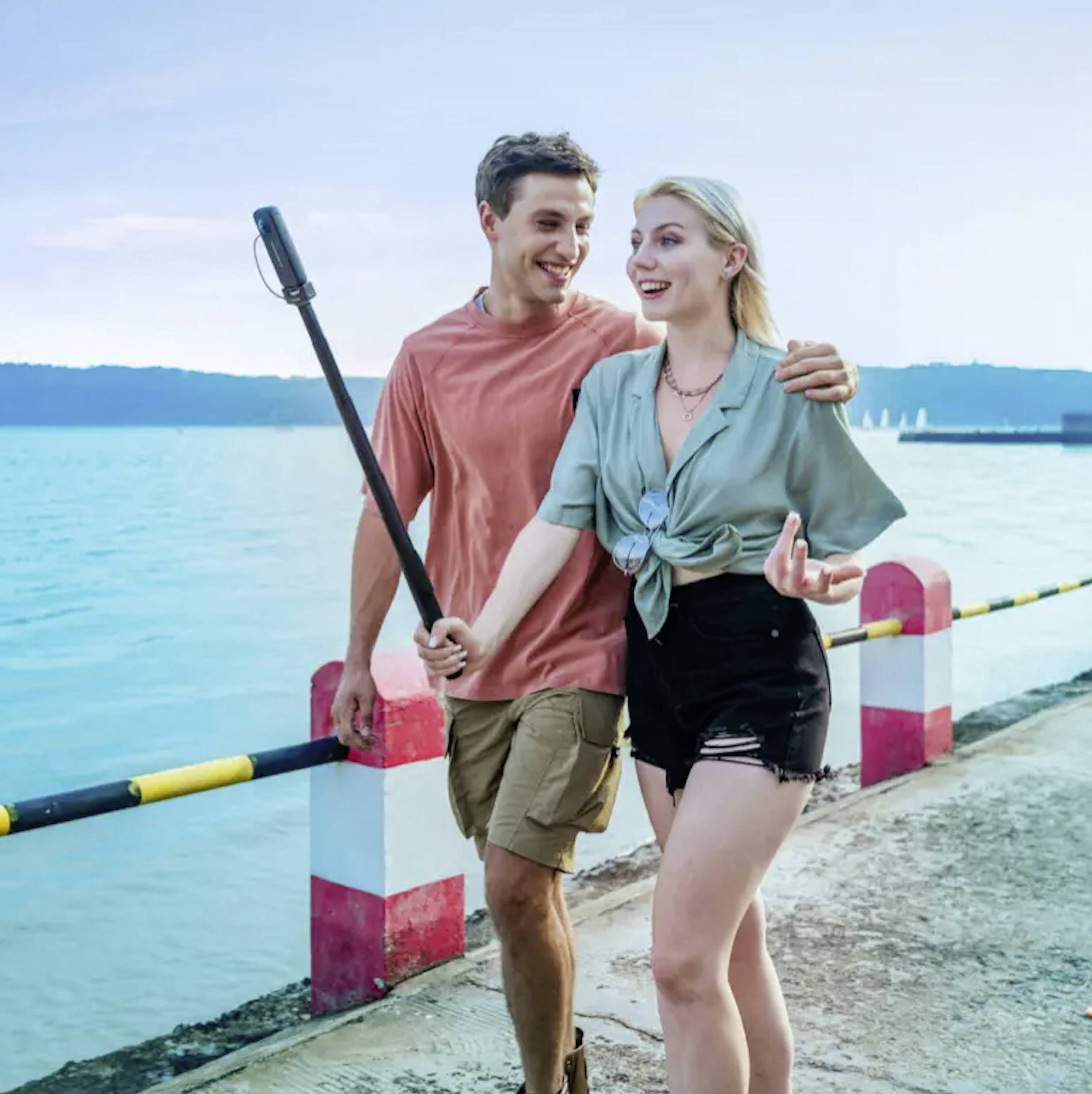 Een lachend stel loopt langs een boulevard en maakt een selfie met een selfie-stick. Op de achtergrond is de zee zichtbaar.