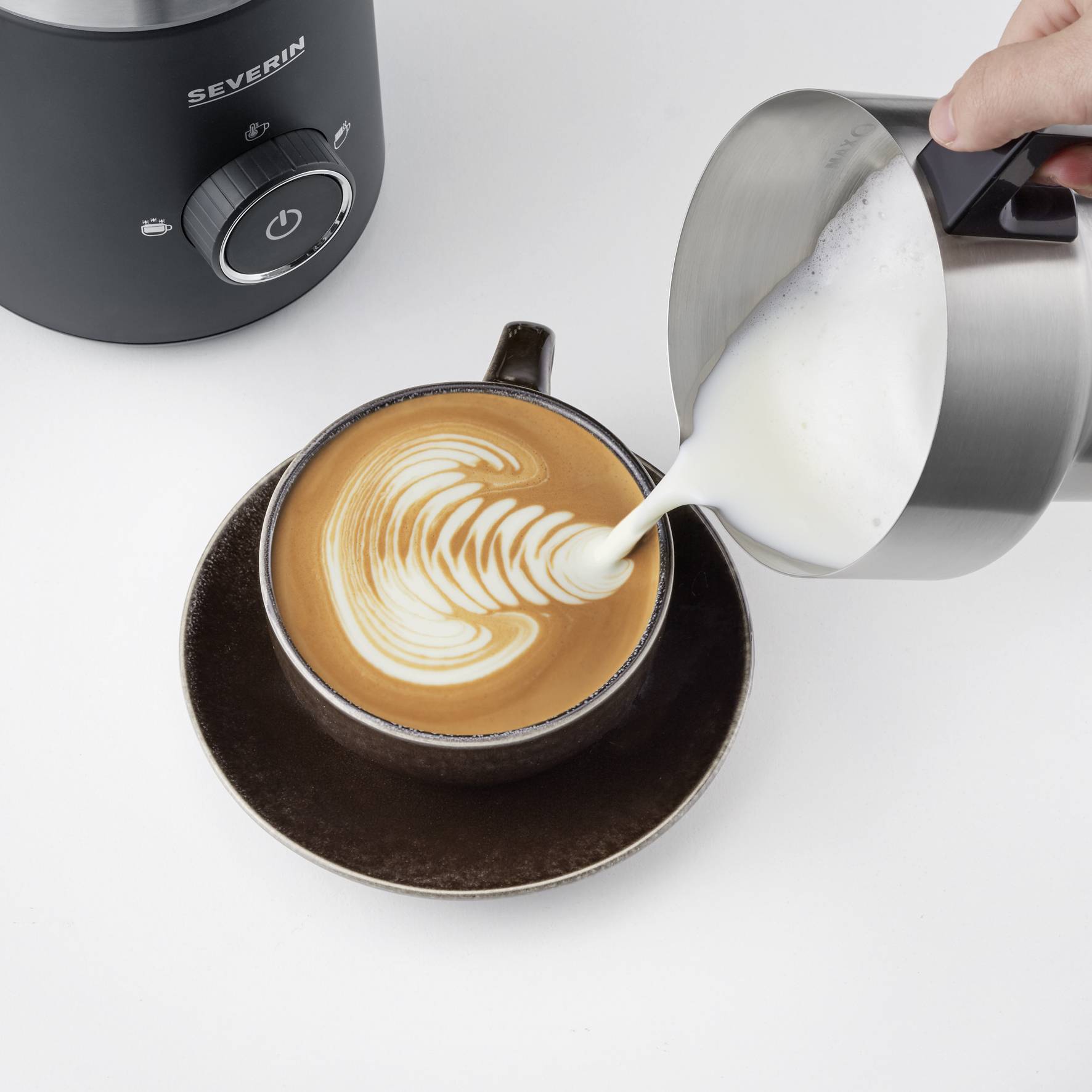 'Een bruine mok met latte art wordt gevuld met melk uit een metalen kan. Op de achtergrond is een melkopschuimer zichtbaar.'