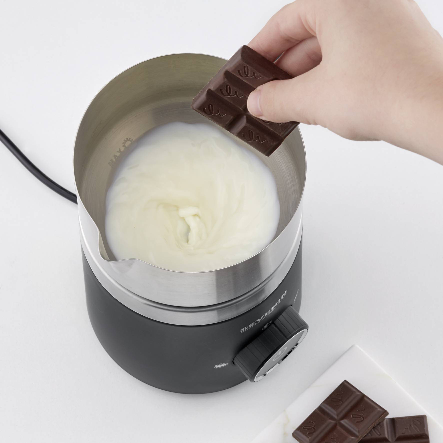 Een hand houdt een stuk chocolade boven een pan met draaiende melkroom in een elektrische mixer.