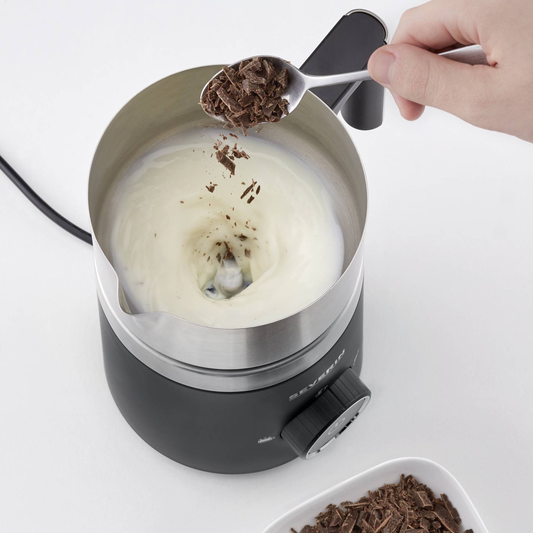 'Kom van een elektrische chocolade-fondue. Een hand houdt een lepel met geraspte chocolade boven de verwarmde vloeistof.'