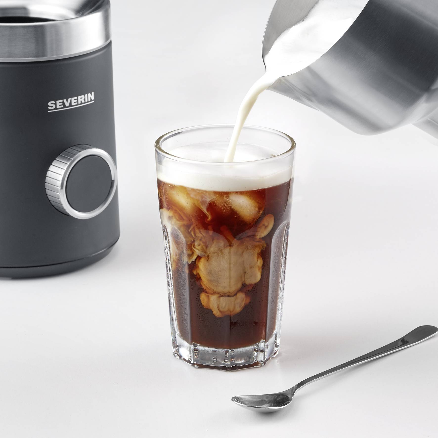 Een glas ijskoffie waarop melk wordt gegoten, naast een zwarte melkopschuimer en een theelepel.