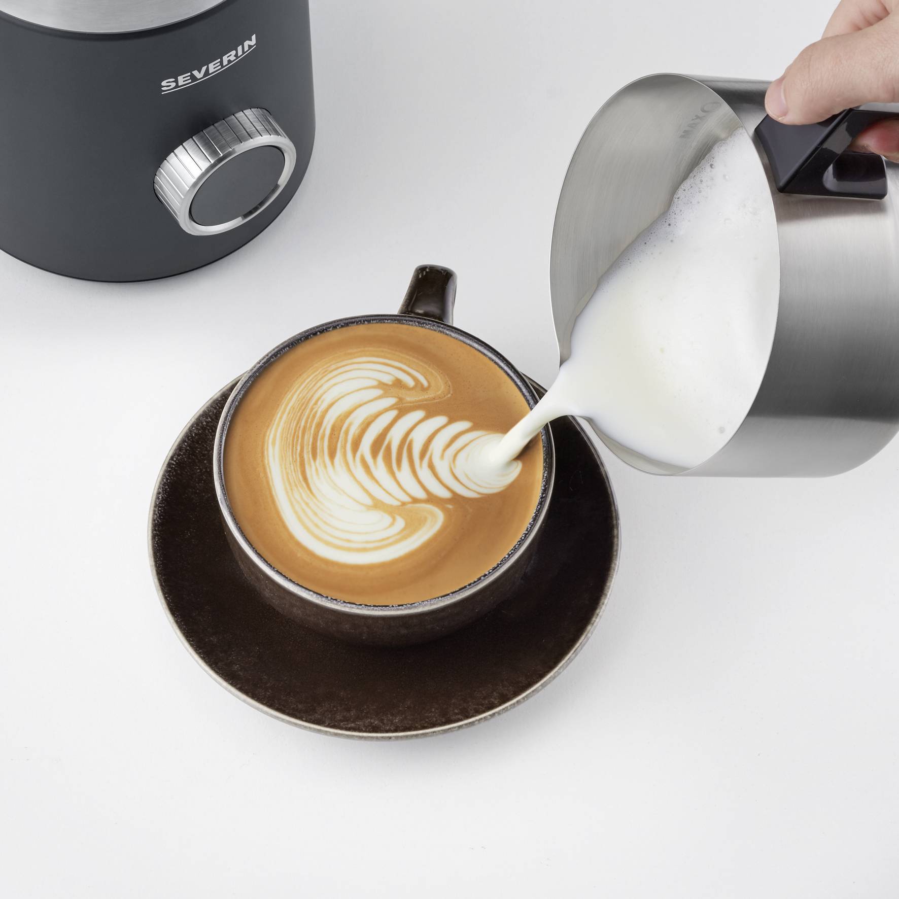 'Een cappuccino met latte art wordt door een hand in een kopje geschonken, naast een elektrische melkopschuimer.'