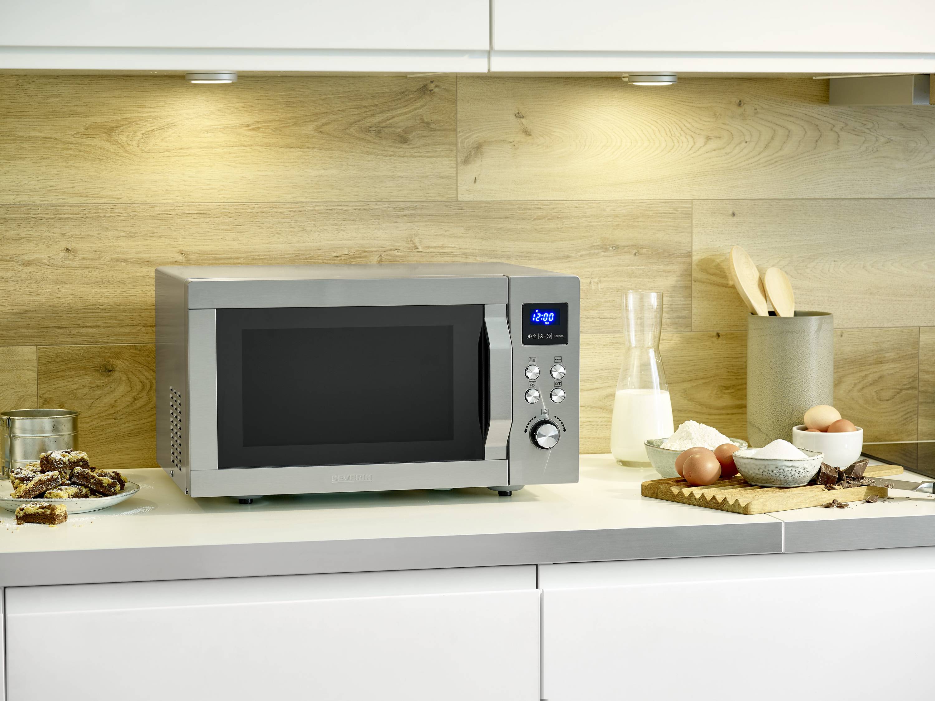 Een zilveren magnetron staat in een moderne keuken. Rechts ernaast bevinden zich een melkfles, eieren en een snijplank.