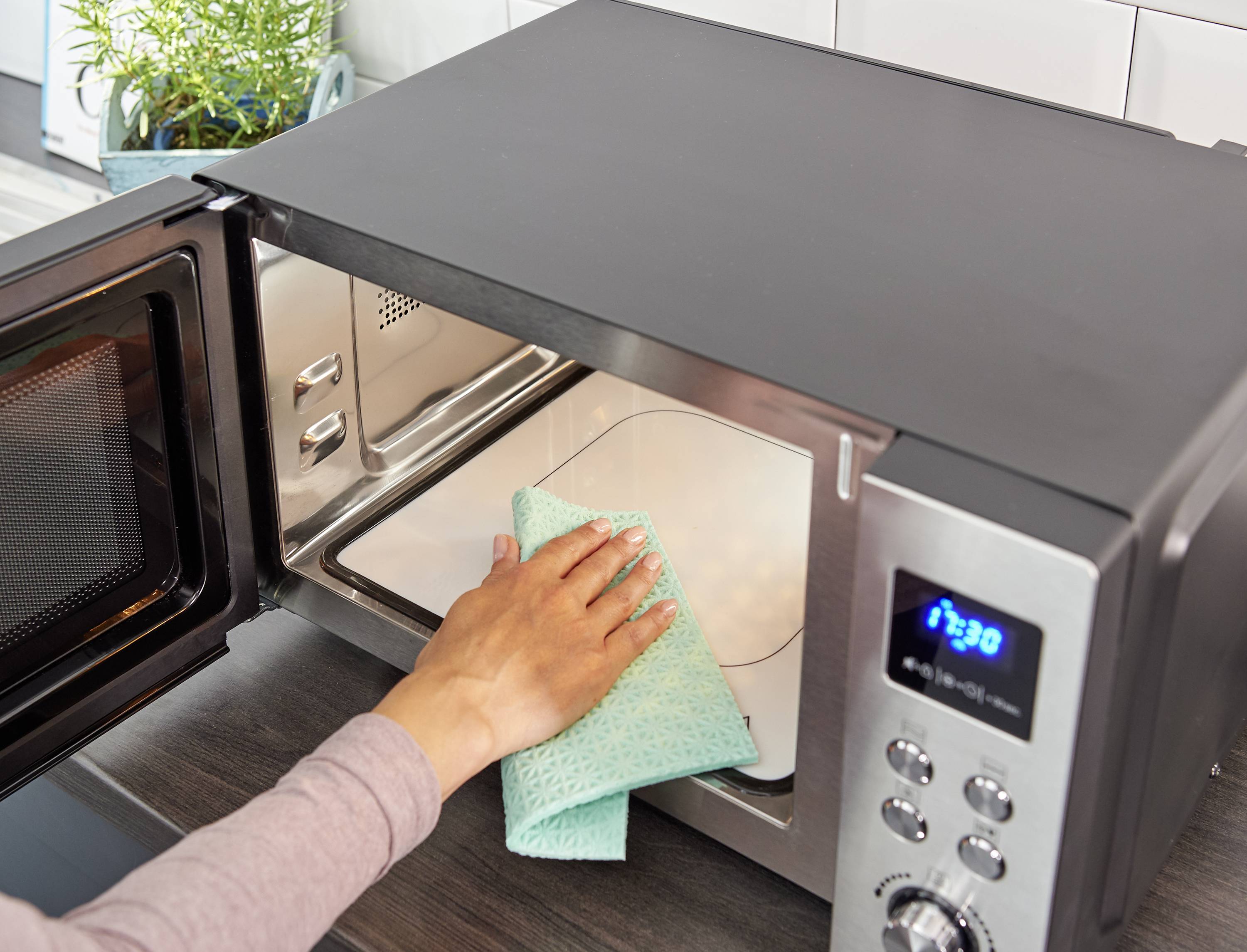 Persoon reinigt de binnenkant van een magnetron met een doek. De magnetron staat op een keukentafel.