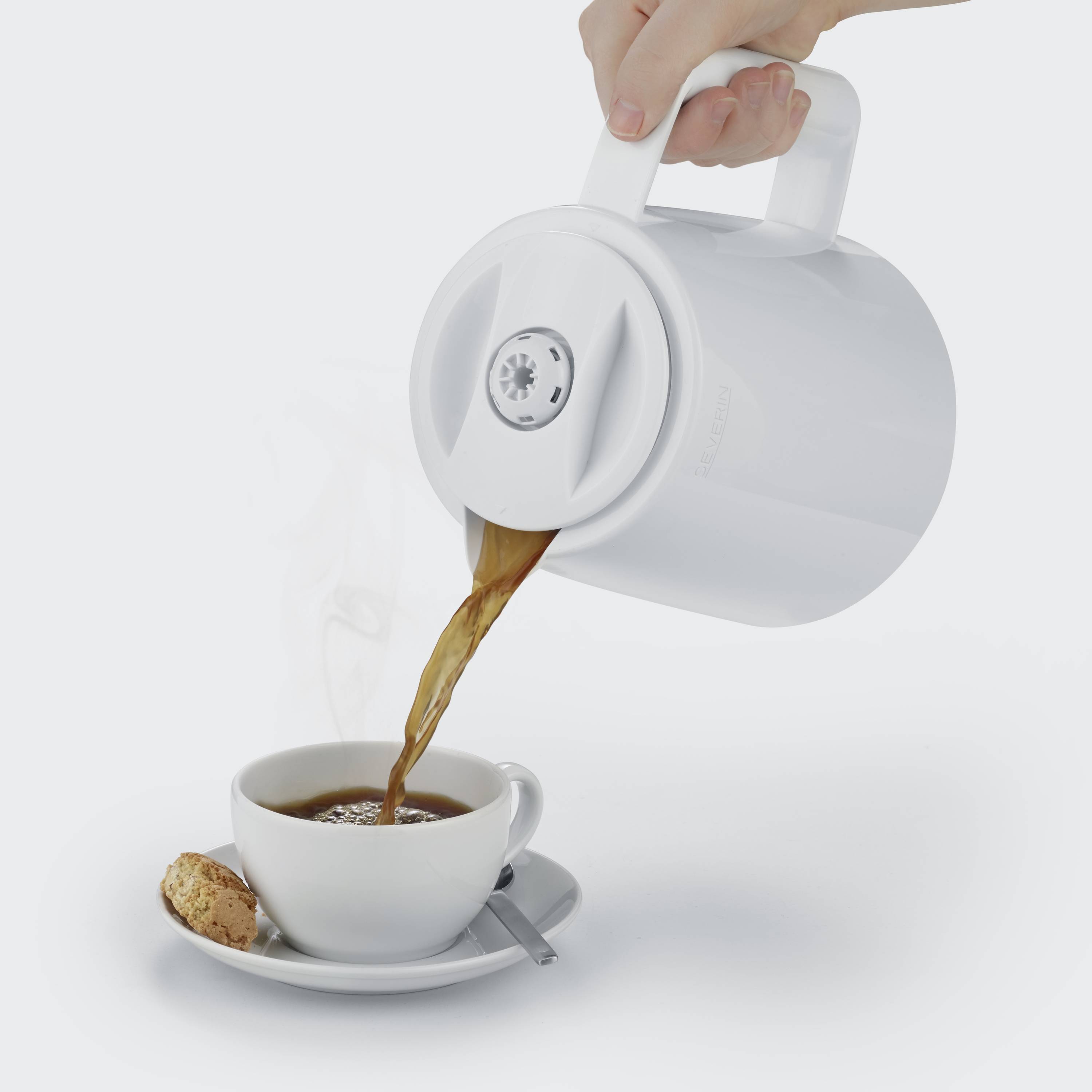 Een witte waterkoker giet hete thee in een kopje met schotel, ernaast ligt een koekje. Stoom stijgt op uit de kop.