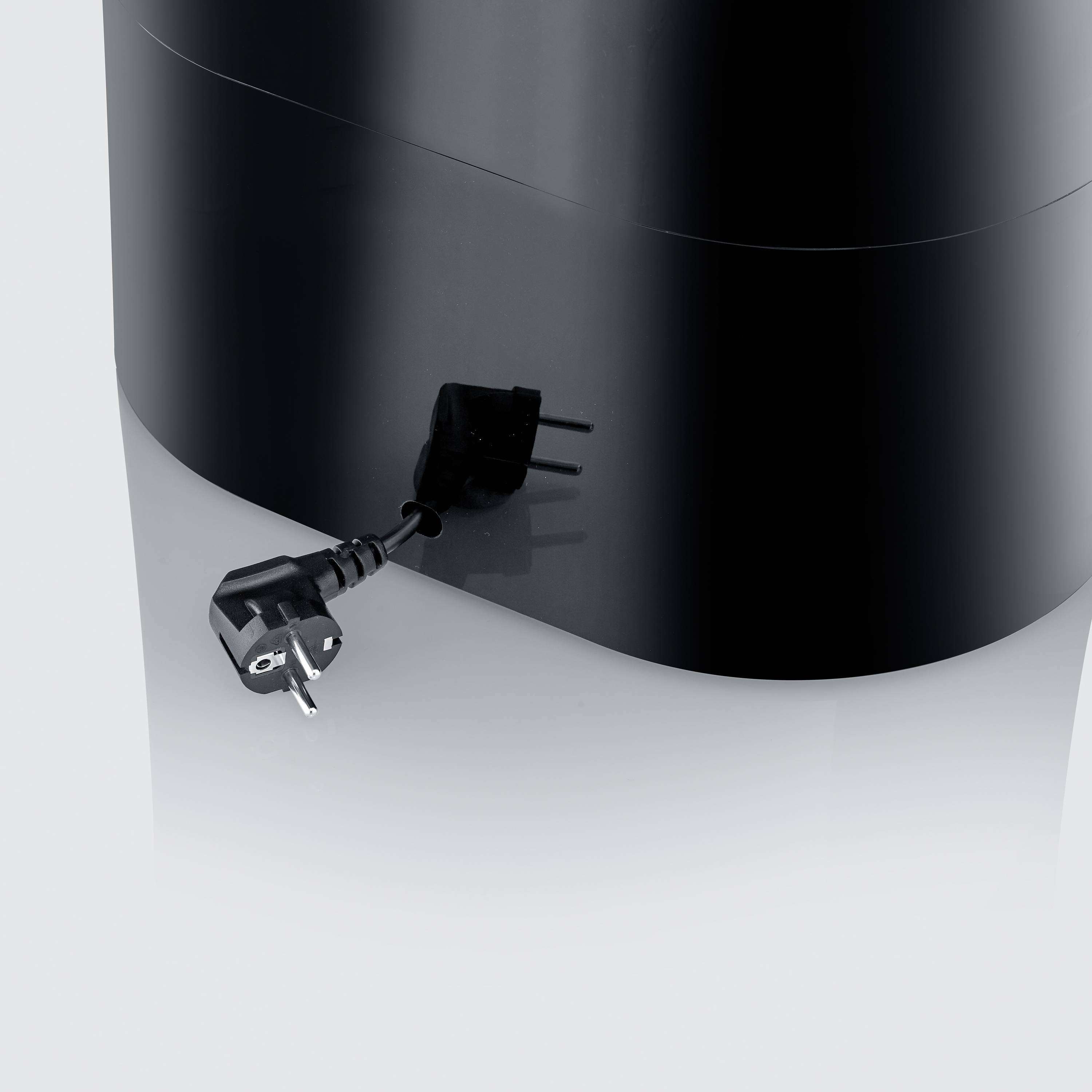 Een zwarte elektrische stekker en een zwart apparaat staan op een reflecterende oppervlakte. De stekker is niet aangesloten.