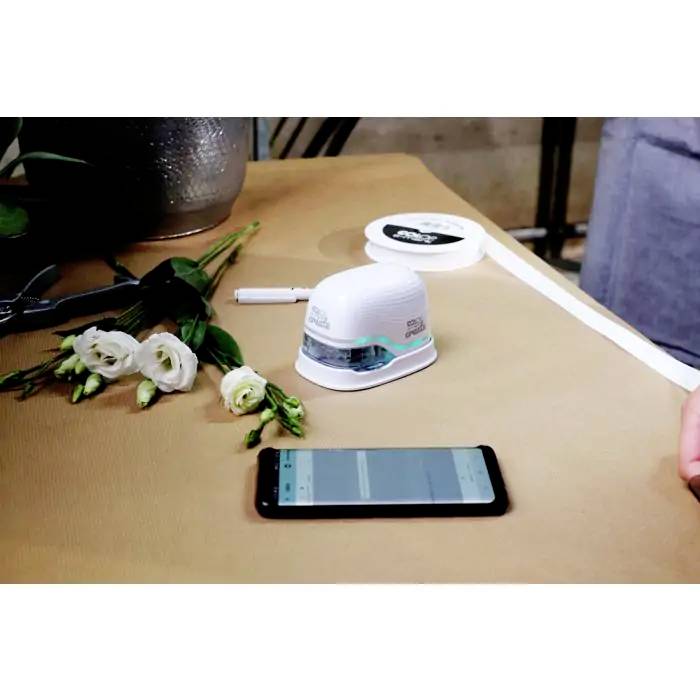 Een kleine printer op een tafel naast een smartphone, bloemen en linten. De printer staat aan en is klaar voor gebruik.