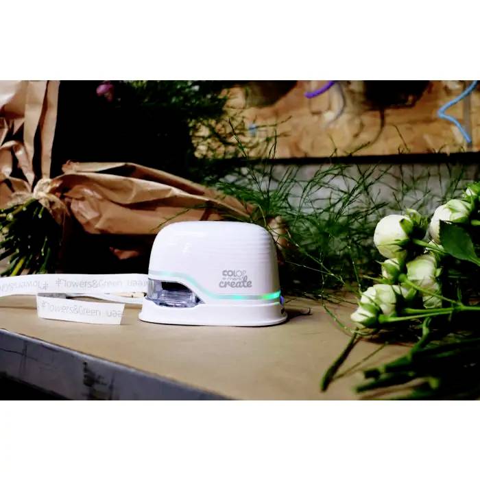 Een kleine, witte printer op een tafel, omgeven door bloemboeketten en groene planten. Op de printer staat de tekst 'COLOP create'.
