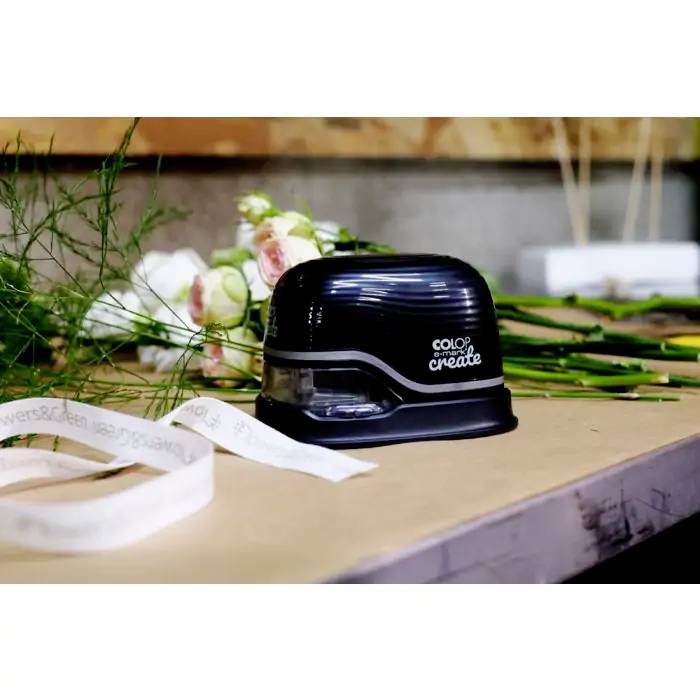 Een etikettenpers met het opschrift 'Colop e-mark create' op een tafel, omringd door bloemen en plantenmaterialen.