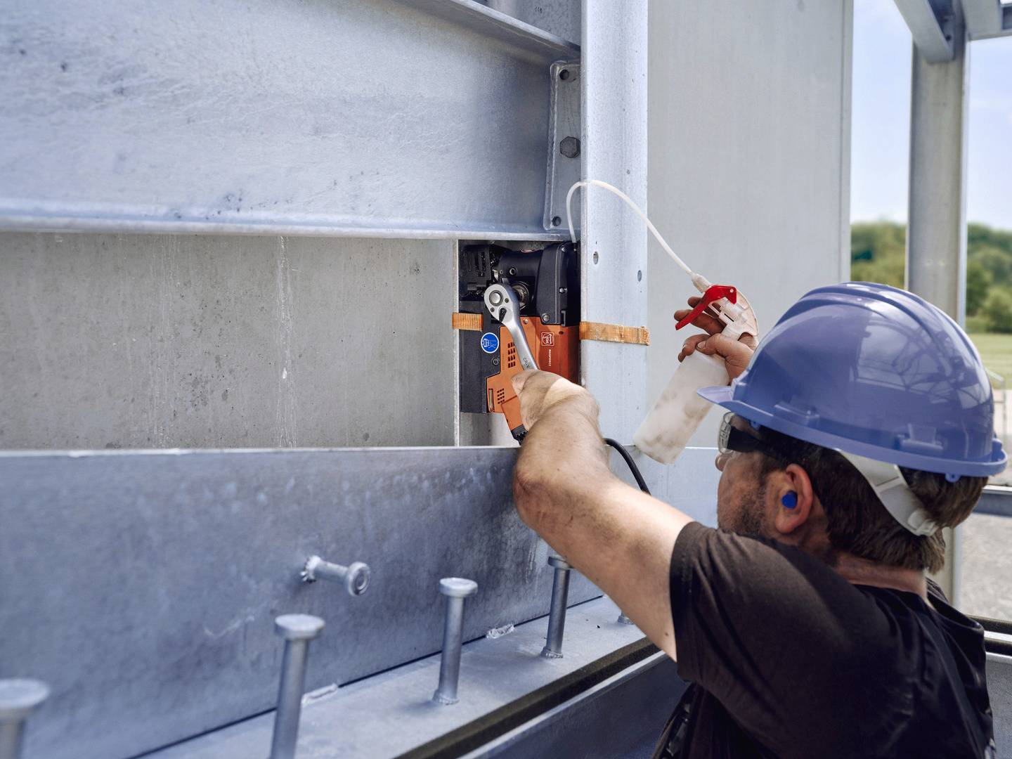 Een arbeider met een veiligheidshelm en oordoppen gebruikt een tang om te werken aan een grote metalen constructie.