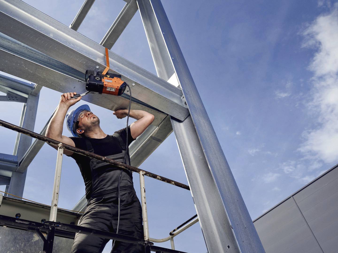 Een bouwvakker op een hoogwerker bevestigt met een gereedschap een ligger aan een staalconstructie onder een blauwe lucht.