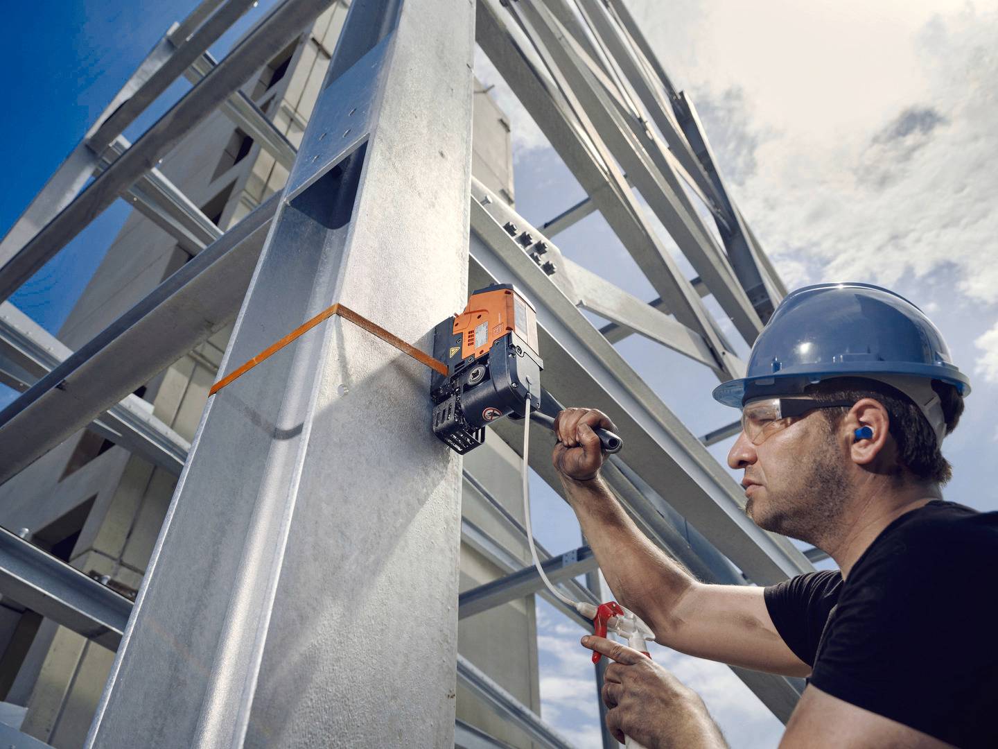 Een arbeider draagt een blauwe veiligheidshelm en bevestigt met een gereedschap een apparaat aan een staalconstructie in de buitenlucht.