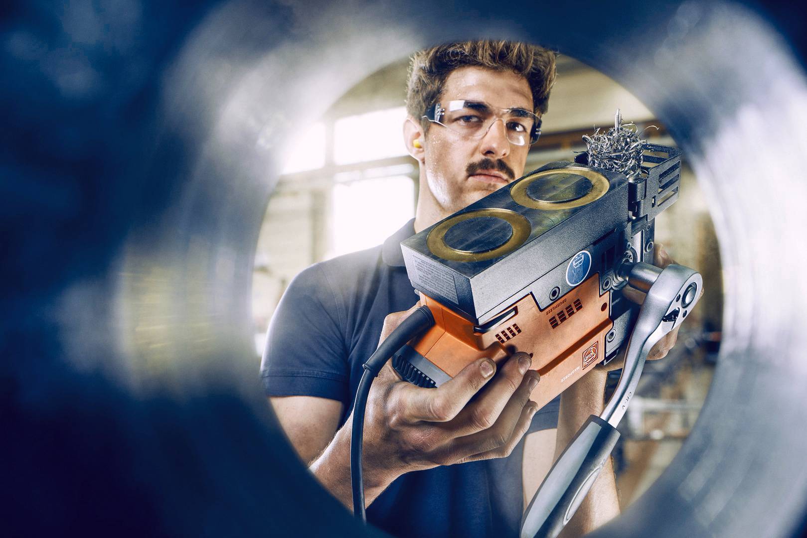 Een man met veiligheidsbril houdt een oranje-zwart apparaat vast, mogelijk een technisch gereedschap. Hij bevindt zich in een industriële omgeving.