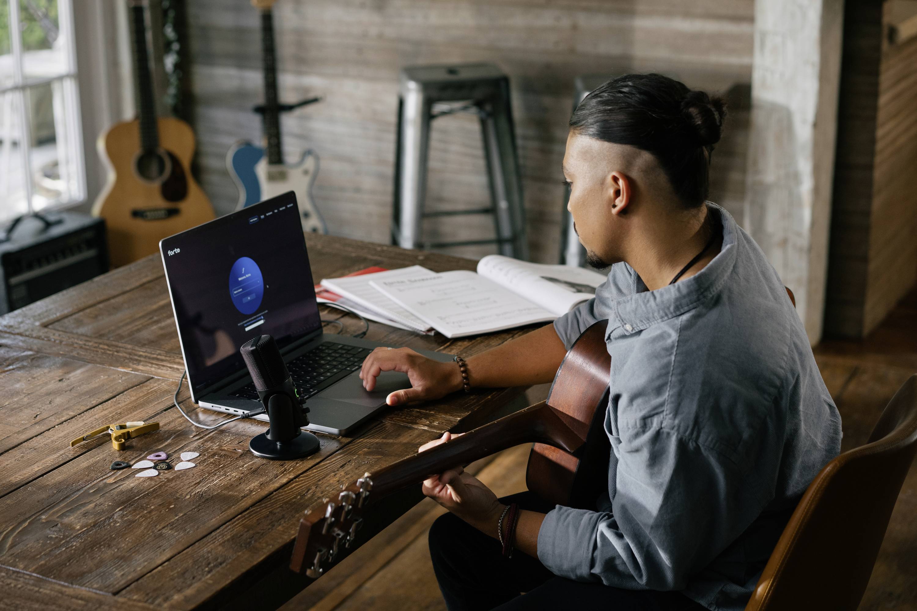 Een persoon zit aan een tafel met een gitaar en werkt op een laptop. Een microfoon en notitieboek liggen ernaast.