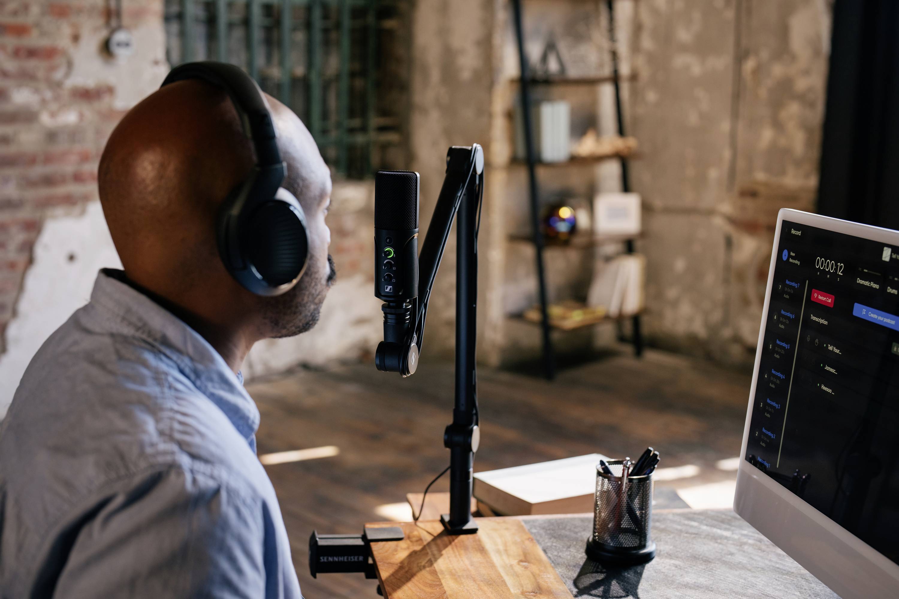 Een persoon met koptelefoon spreekt in een microfoon in een studio. Een computerscherm toont audiosoftware.