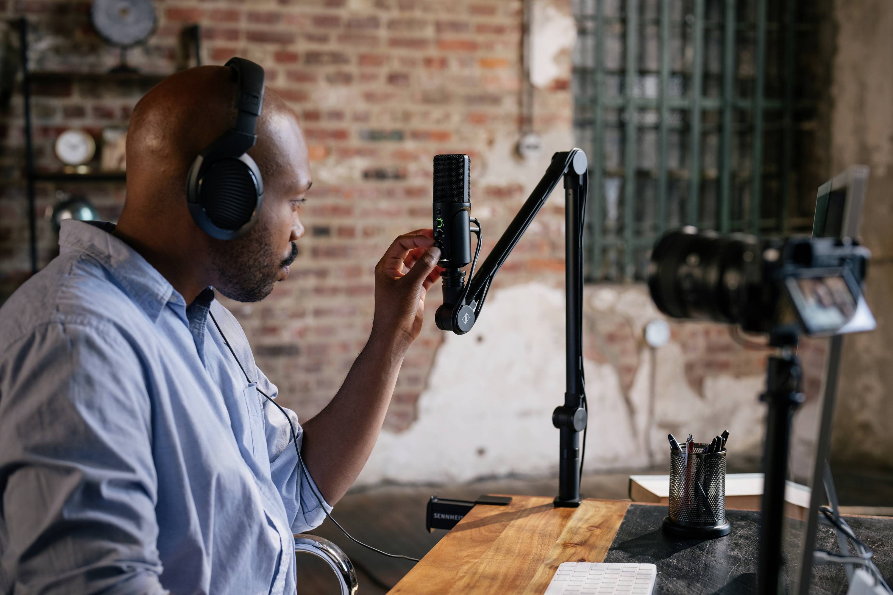 Een man met koptelefoon zit in een studio en stelt een microfoon af. Op de achtergrond zijn bakstenen muren en technische apparatuur zichtbaar.
