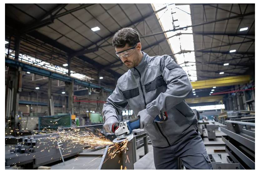 Een man in veiligheidsuitrusting gebruikt een haakse slijper op metaal in een grote industriële werkplaats, waarbij vonken spatten bij het contact.