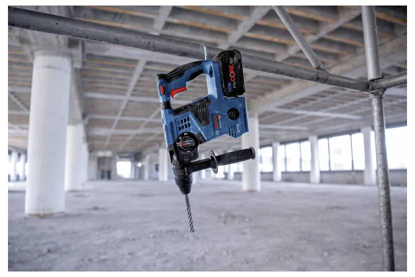 Een blauwe snoerloze klopboormachine met een zwarte handgreep en rode accenten hangt aan een haak in een onafgewerkt betonnen gebouw, waarbij constructiegereedschappen worden geaccentueerd.
