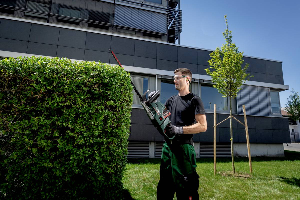 Een man knipt met een elektrische heggenschaar een haag voor een modern gebouw. Hij draagt beschermende handschoenen en een veiligheidsbril.