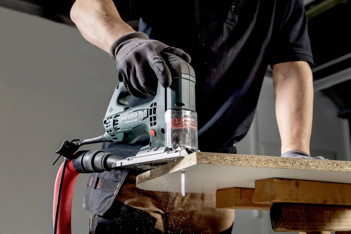 Een persoon zaagt een houten plank met een elektrische decoupeerzaag. Houtkrullen vallen terwijl de zaag door het hout gaat.