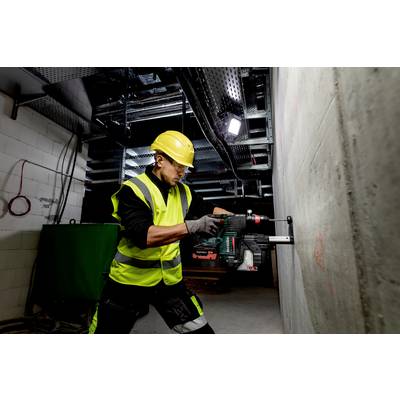Een bouwvakker in veiligheidskleding en helm boort met een boormachine in een betonnen muur in een industriële ruimte.