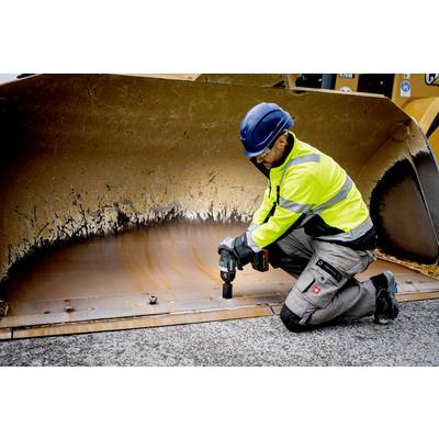 Een bouwvakker in beschermende werkkleding knielt voor een grote graafmachinebak en werkt met een boormachine aan de rand van de bak.
