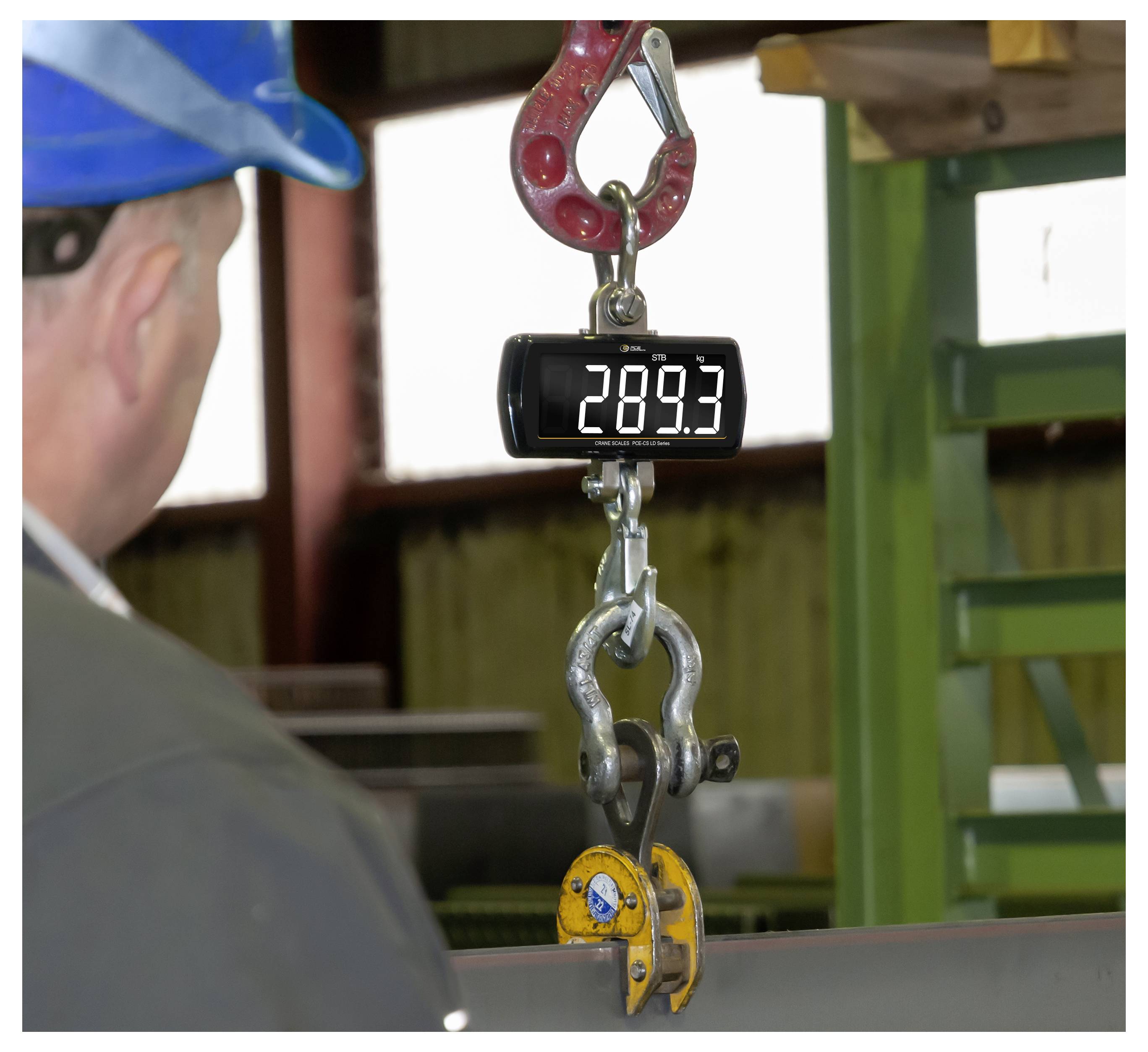 Een digitale hangweegschaal toont 289,3 terwijl een werker in een veiligheidhelm een opgehangen metalen haak en klem in een industriële omgeving in de gaten houdt.
