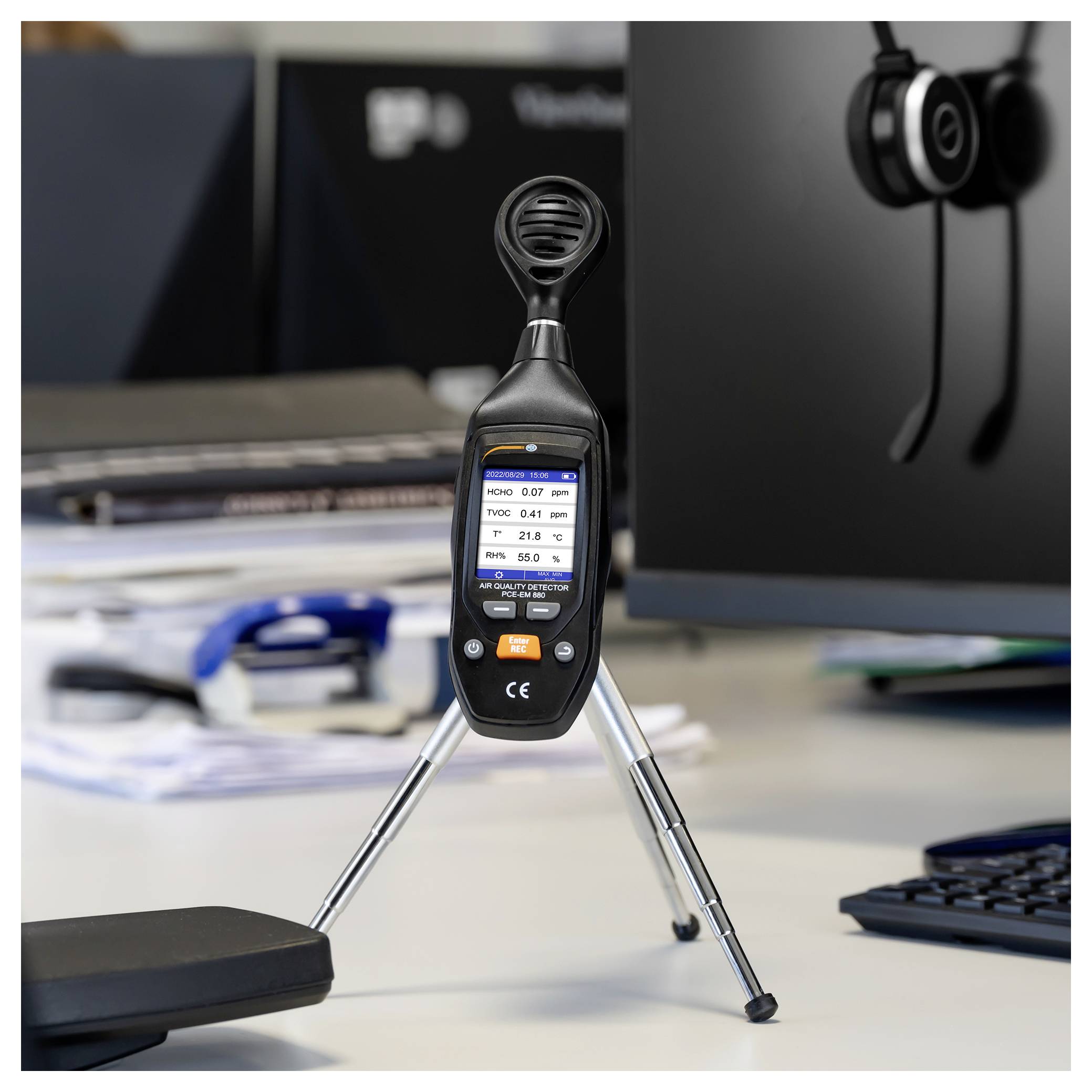 Een handheld luchtkwaliteitsmeter op een statief toont meetwaarden op zijn scherm in een kantooromgeving aan een bureau.