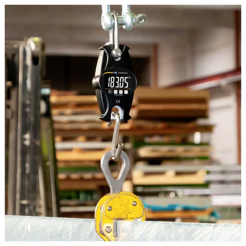 Digitale hangweegschaal bevestigd aan een metalen haak, met een weergave van 183.05, hangend boven een gele hefklem in een industriële omgeving.
