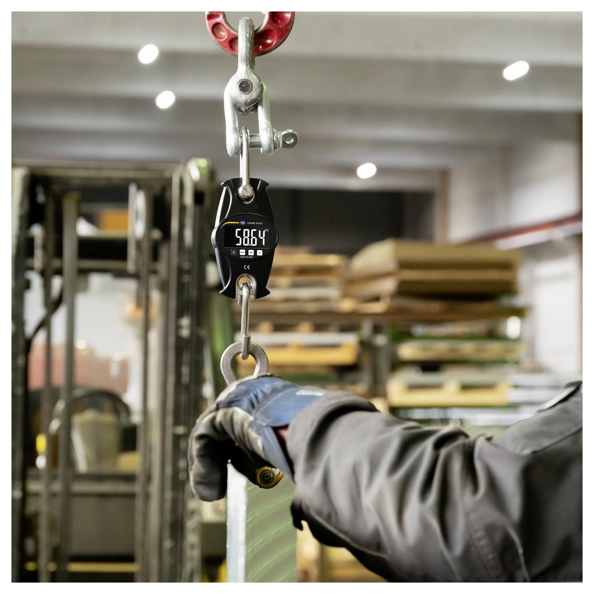 Gehandschoende werker die een hangende lading verwerkt die aan een digitale haaklood is bevestigd en 5864 aangeeft, in een industrieel magazijn.