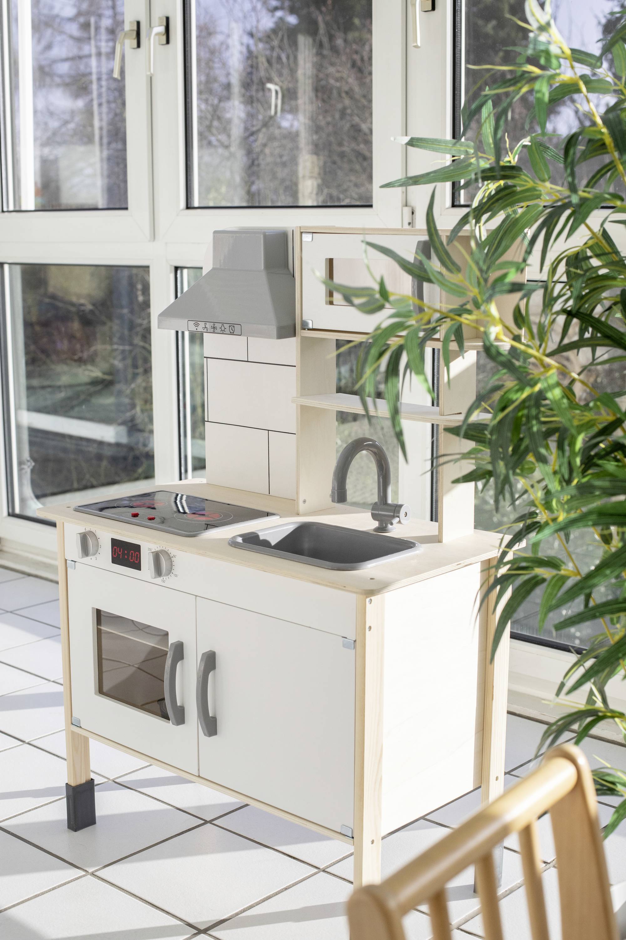 Kinderkeuken met fornuis, gootsteen en oven van hout staat in een zonnige ruimte nabij een groot raam. Ernaast staat een plant.