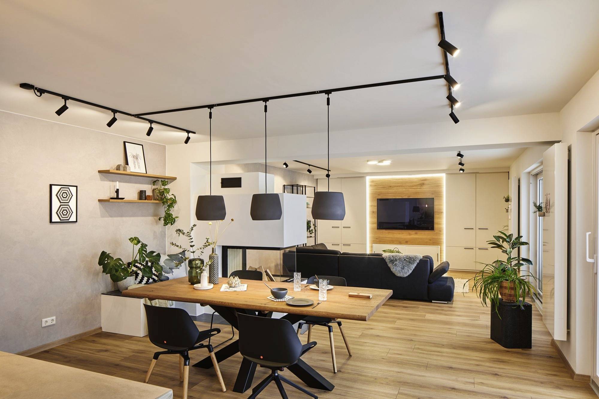 Een modern woonvertrek met eettafel, hangende lampen en bank, omringd door planten en decoratieve boekenplanken, in warme houtachtige tinten.