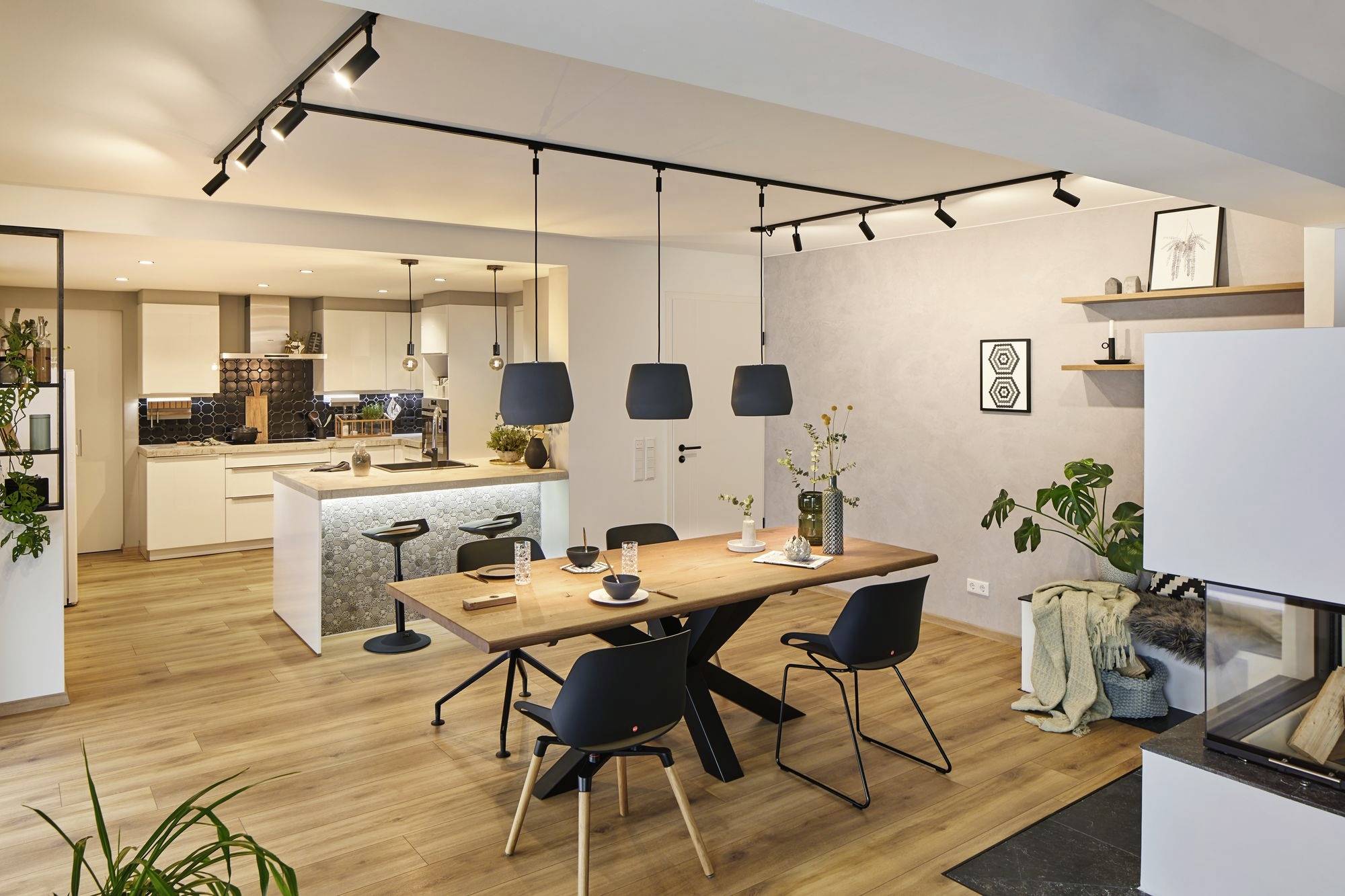 Moderne eetkamer met houten tafel en vier zwarte stoelen onder drie hangende lampen. Op de achtergrond een open keuken met een keukeneiland.